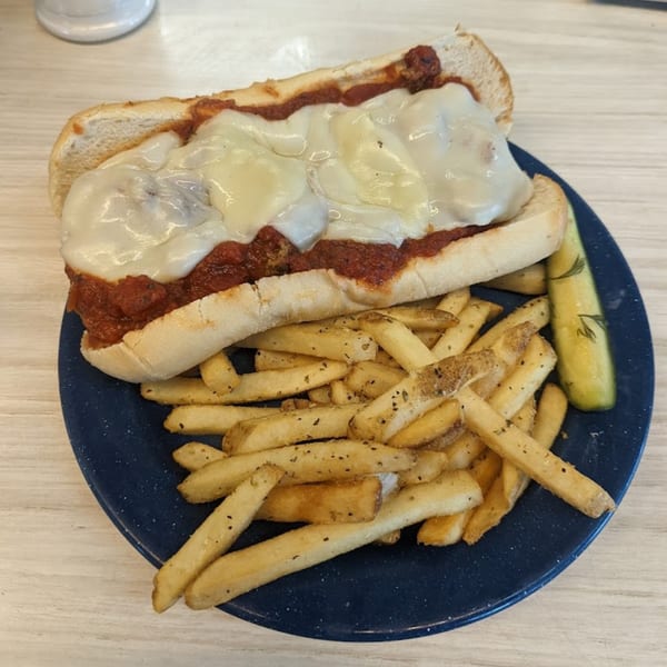 A meatball sub with melted cheese and marinara sauce, served with fries and a pickle spear.